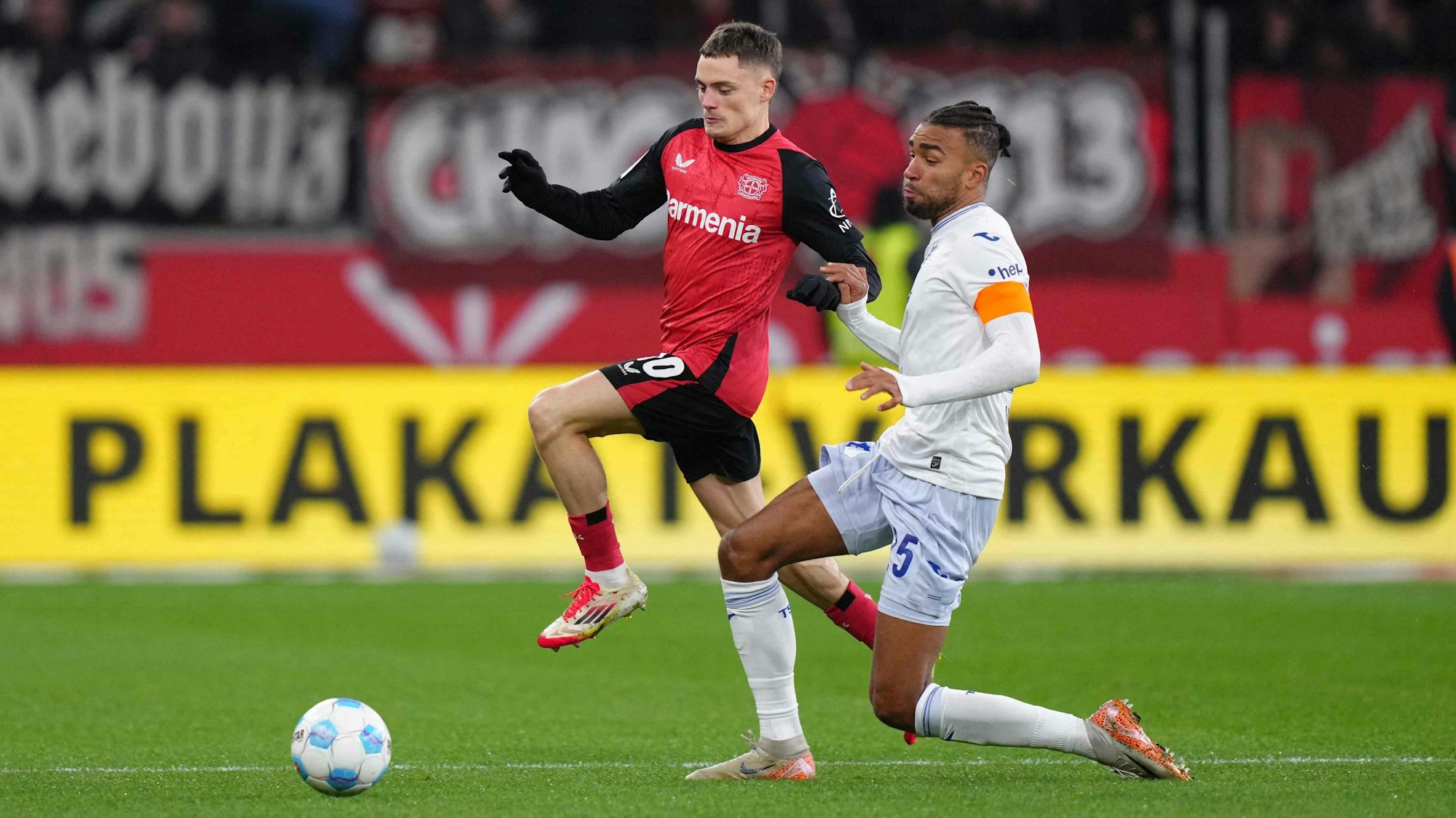 Das Bild zeigt einen Zweikampf zwischen Florian Wirtz und Kevin Akpoguma.