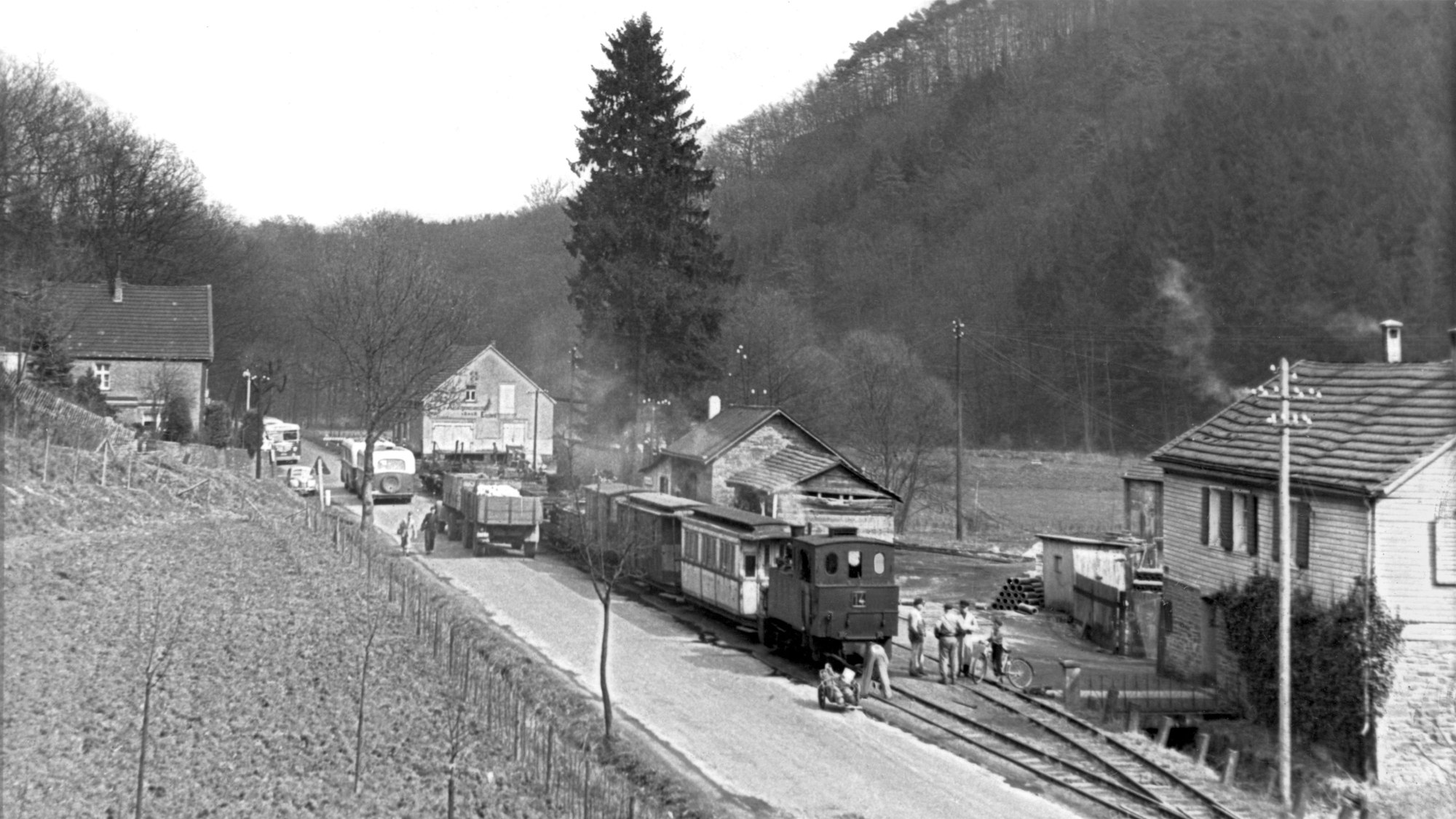 Ein Foto von 1954: Eine Schmalspureisenbahn auf einer einsamen Straße, zwei alte Omnibusse mit Anhänger befahren die Straße.