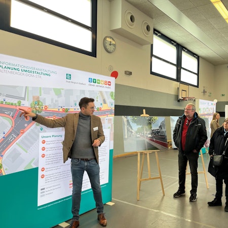 Der Planer steht vor einer Stellwand. Besucher hören ihm zu.