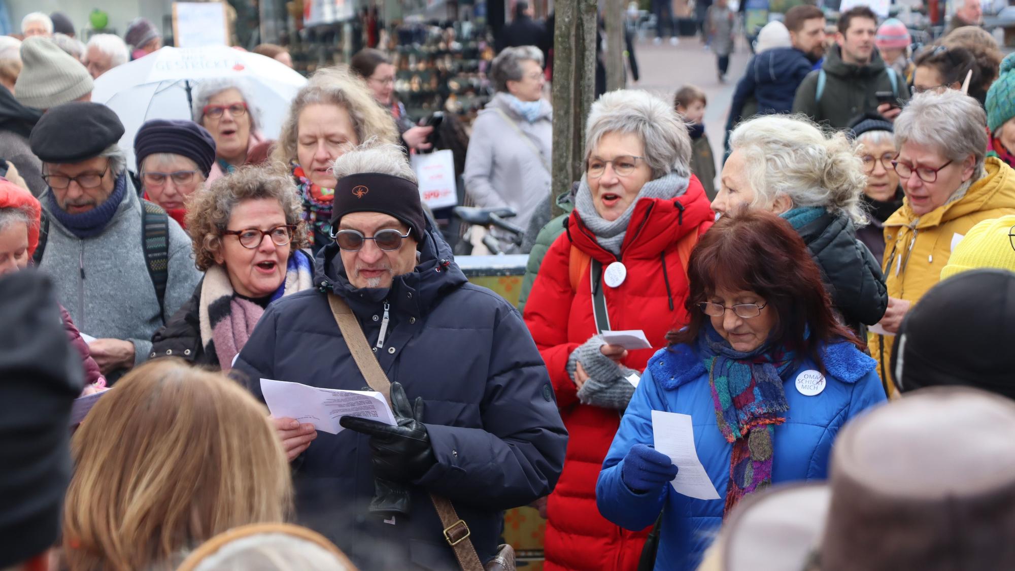 Auf dem Bild ist eine Menschenmenge in der Brühler Fußgängerzone zu sehen. Die Leute halten Zettel mit Liedtexten in der Hand und singen.