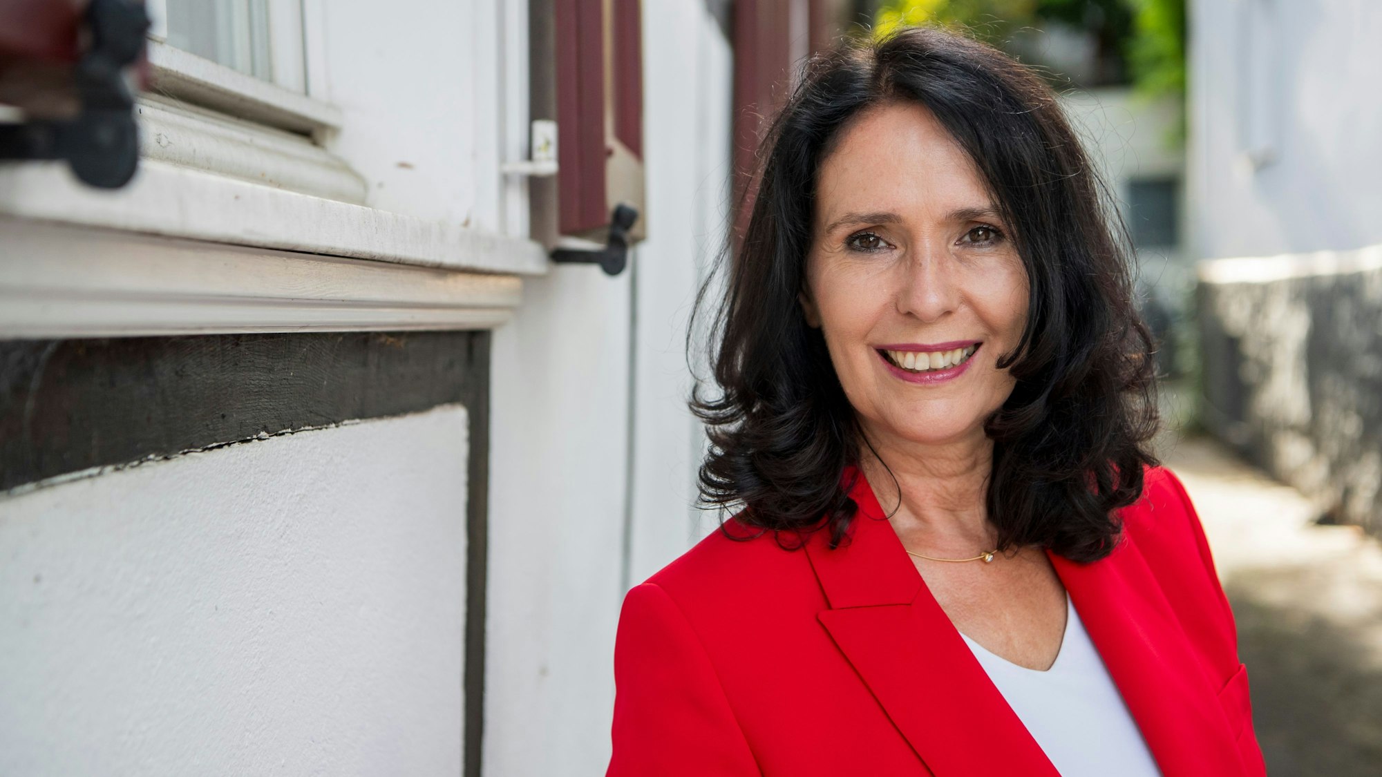 Eine Frau in einem roten Sakko steht an einer Hauswand.