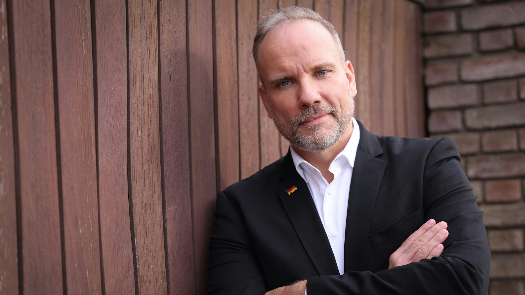 Ein Mann im Anzug lehnt an einer Wand aus Holzbrettern.