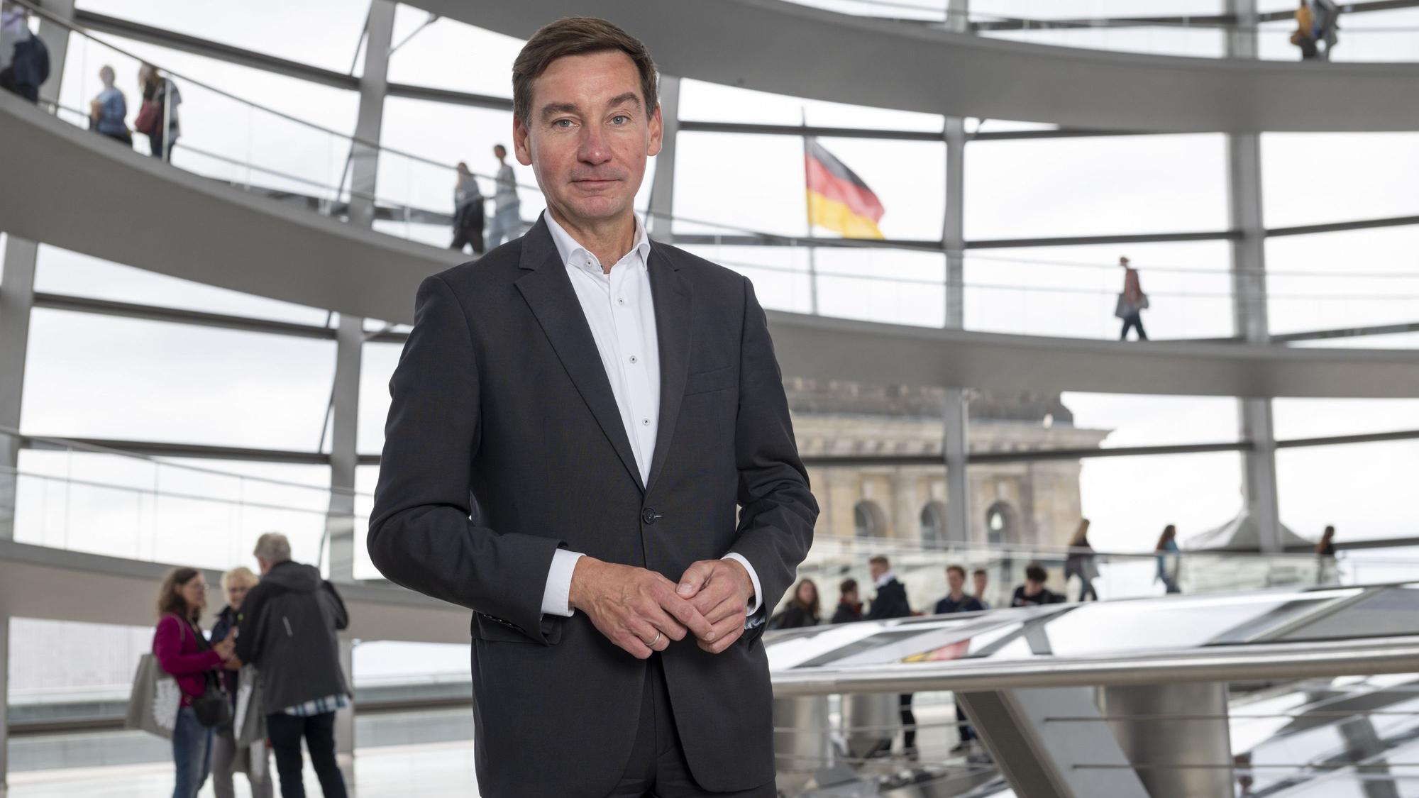 Ein Mann im Anzug steht in einem Gebäude mit großen Fenstern. Im Hintergrund weht eine Deutschlandflagge.