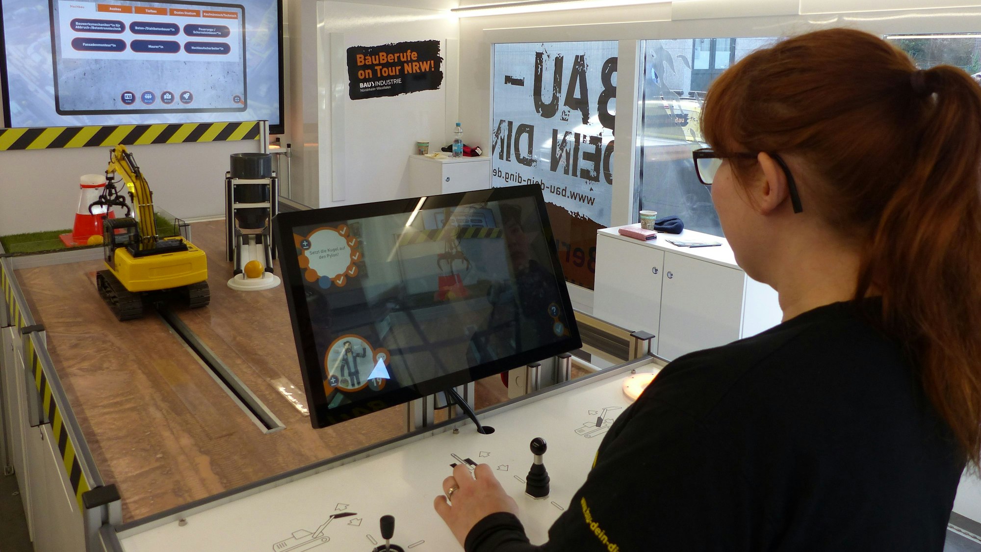 Auf dem Bild sieht man eine Frau, die vor sich einige Hebel und einen Bildschirm hat. Dahinter ein Miniaturbagger.