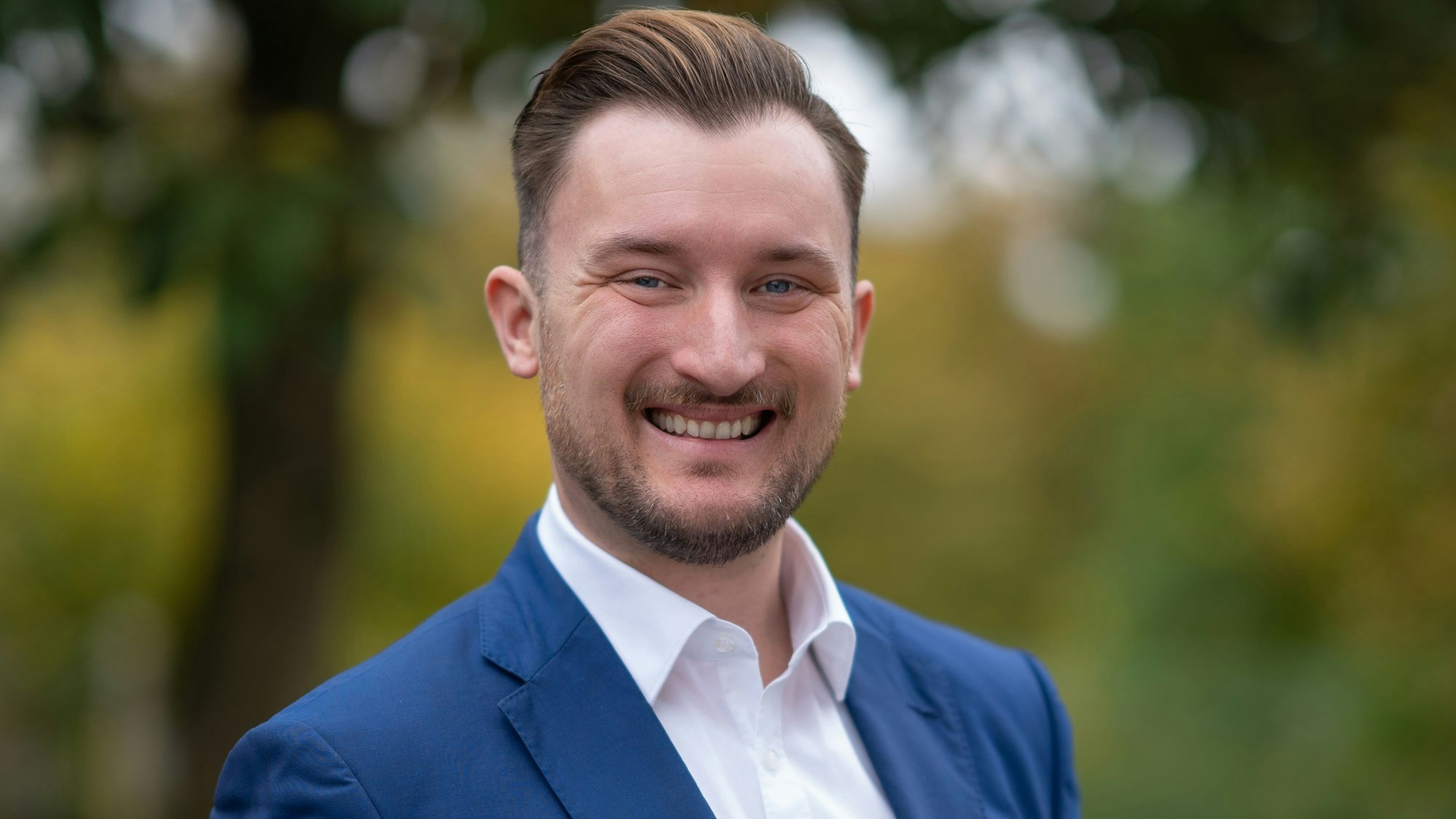 Ein Mann im Anzug steht vor einem unscharfen Hintergrund in der Natur.