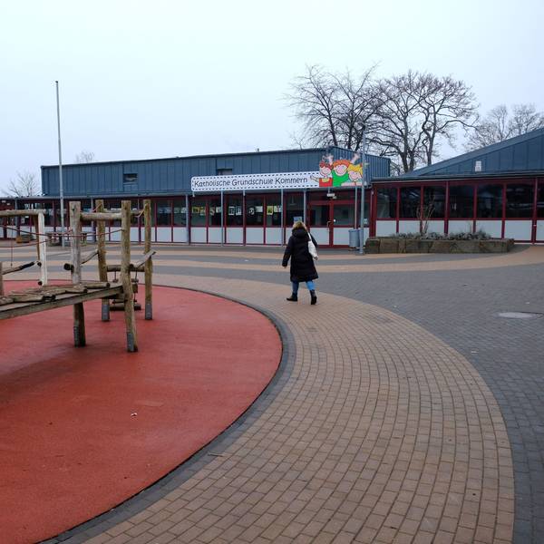 Eine Frau geht über den Pausenhof der Katholischen Grundschule in Kommern. Links im Bild ist ein Klettergerüst aus Holz zu sehen.