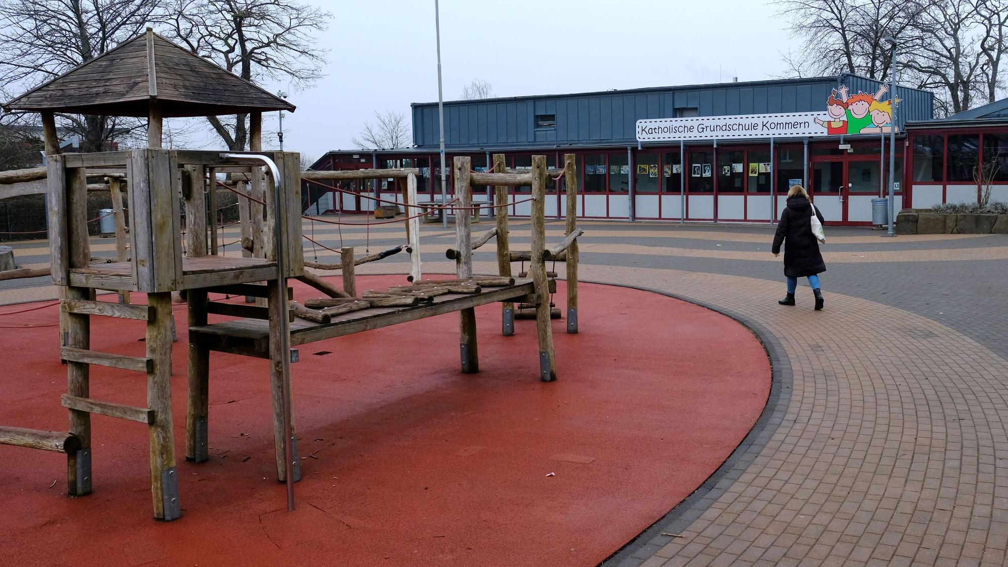 Eine Frau geht über den Pausenhof der Katholischen Grundschule in Kommern. Links im Bild ist ein Klettergerüst aus Holz zu sehen.