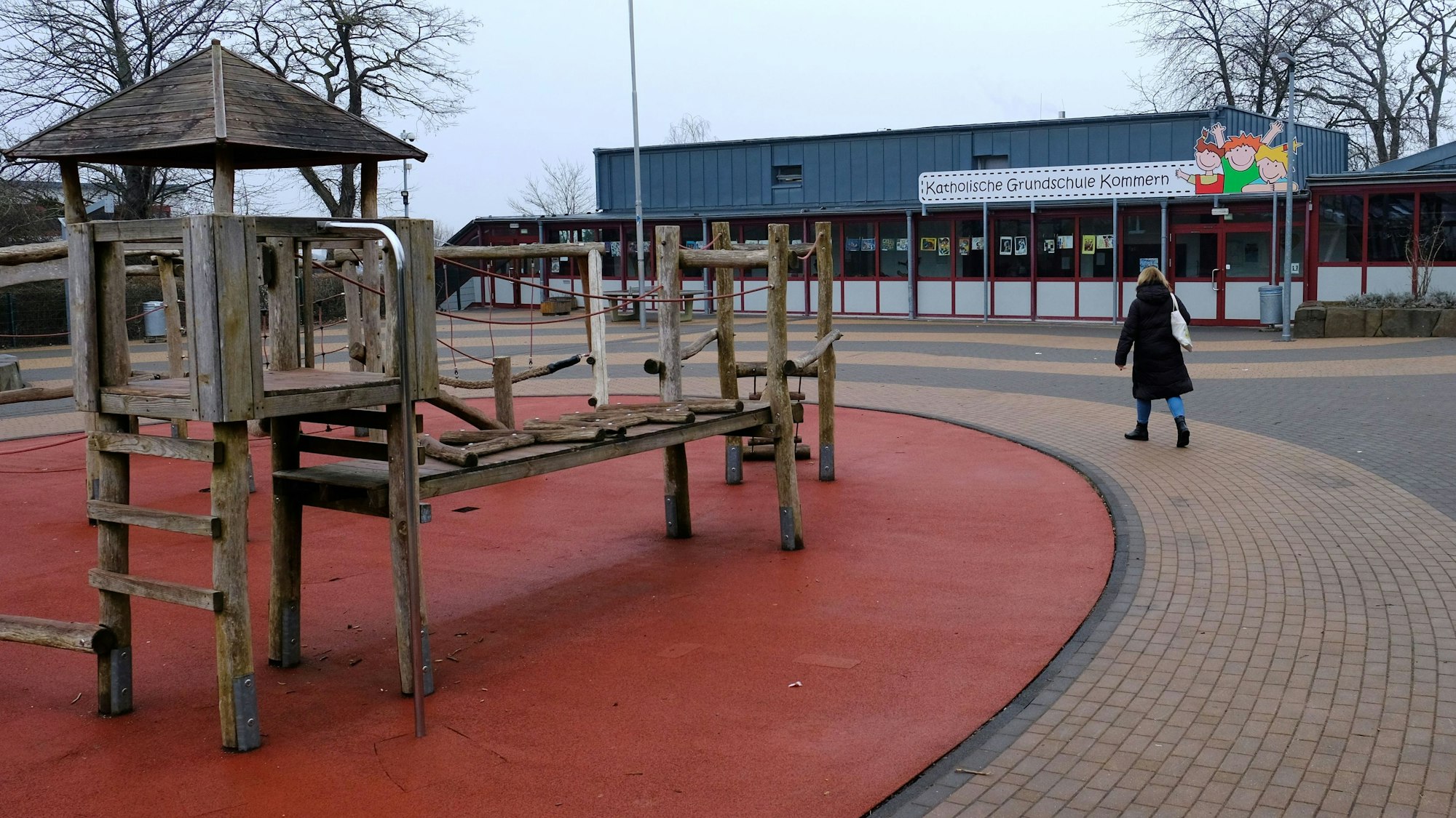 Eine Frau geht über den Pausenhof der Katholischen Grundschule in Kommern. Links im Bild ist ein Klettergerüst aus Holz zu sehen.