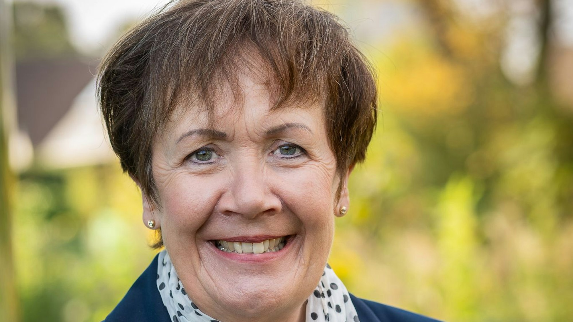 Eine Frau in einem Sakko trägt Schal und steht vor einem unscharfen Natur-Hintergrund.