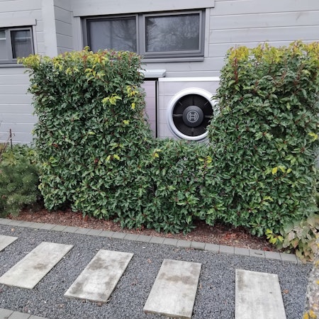 Eine Wärmepumpe vor einem Einfamilienhaus, umrankt von einer immergrünen Hecke