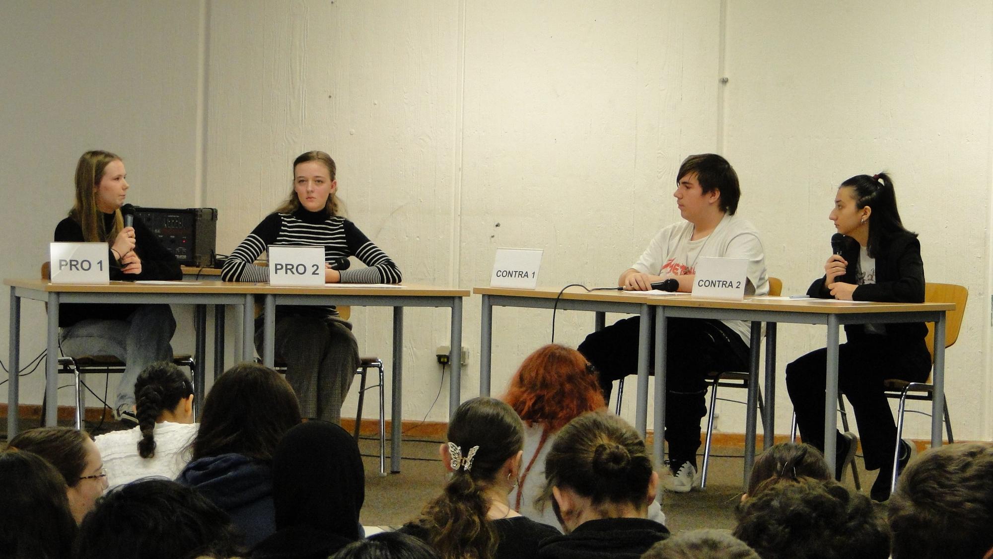 Drei Mädchen und ein Junge sitzen an Schultischen und Debattieren.