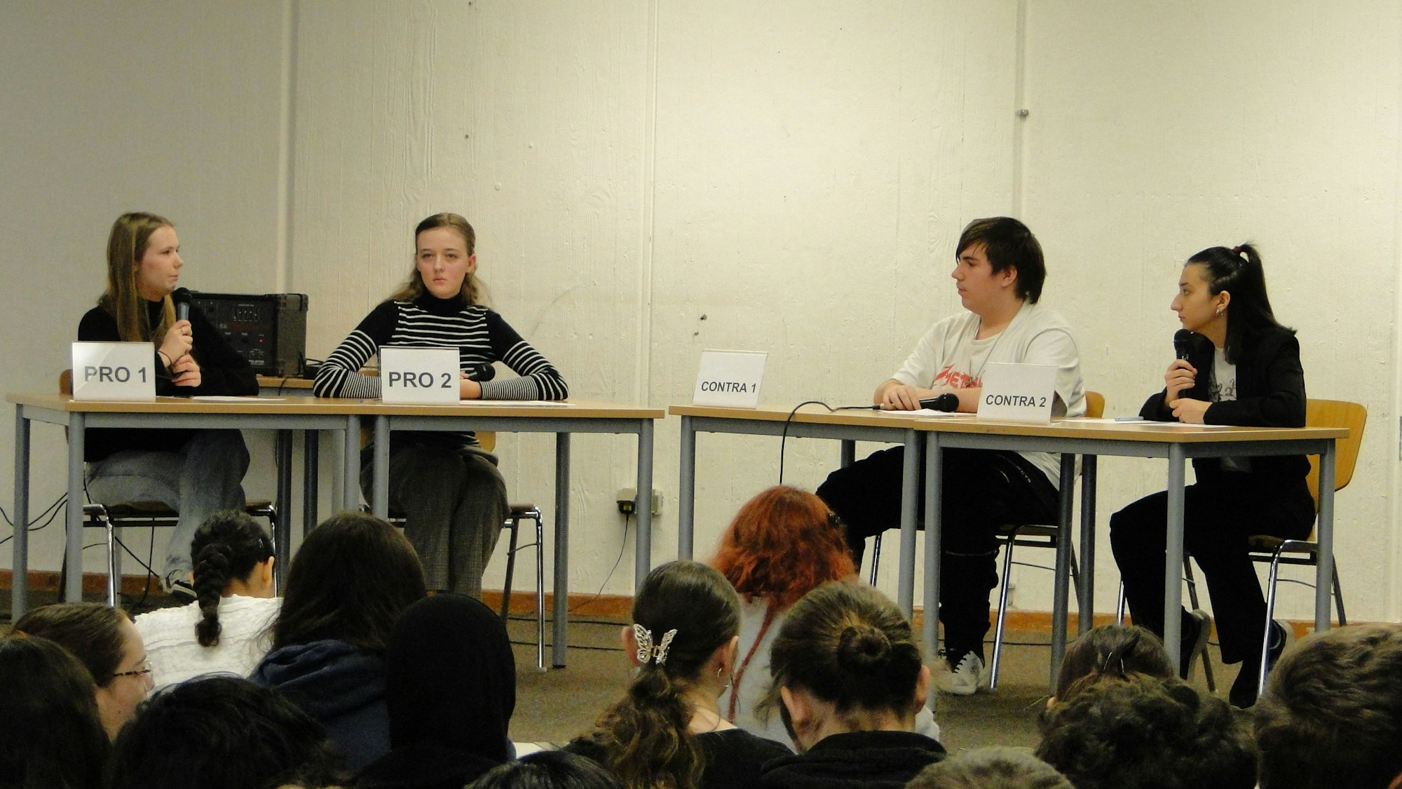 Drei Mädchen und ein Junge sitzen an Schultischen und Debattieren.