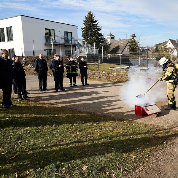 Feuerwehrleute stehen im Rahmen einer Vorführung um eine qualmende Kiste herum. Eine Einsatzkraft unter Atemschutz steht direkt daran und zeigt, wie die Feuerwehr vorgehen kann.