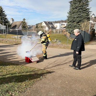 Feuerwehrleute stehen im Rahmen einer Vorführung um eine qualmende Kiste herum. Eine Einsatzkraft unter Atemschutz steht direkt daran und zeigt, wie die Feuerwehr vorgehen kann.