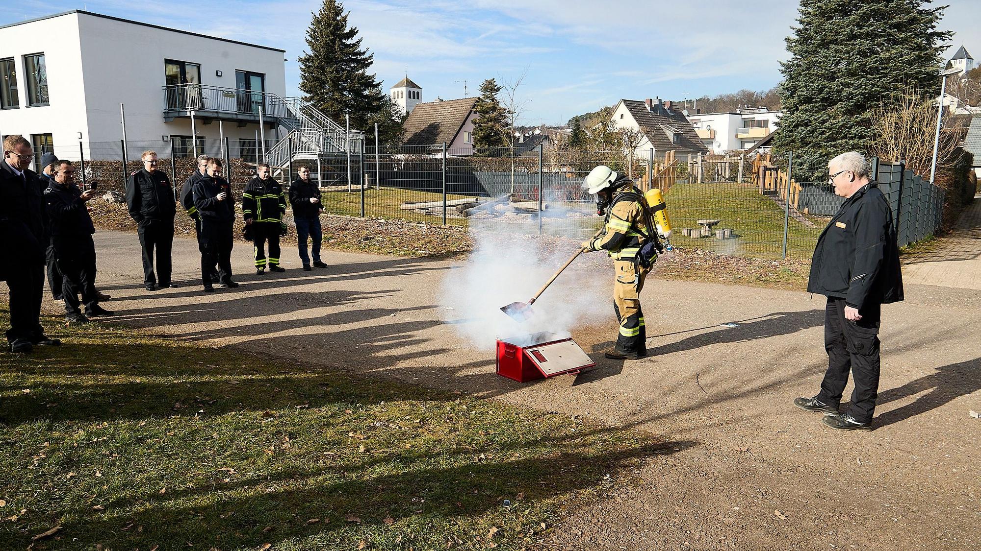 Feuerwehrleute stehen im Rahmen einer Vorführung um eine qualmende Kiste herum. Eine Einsatzkraft unter Atemschutz steht direkt daran und zeigt, wie die Feuerwehr vorgehen kann.