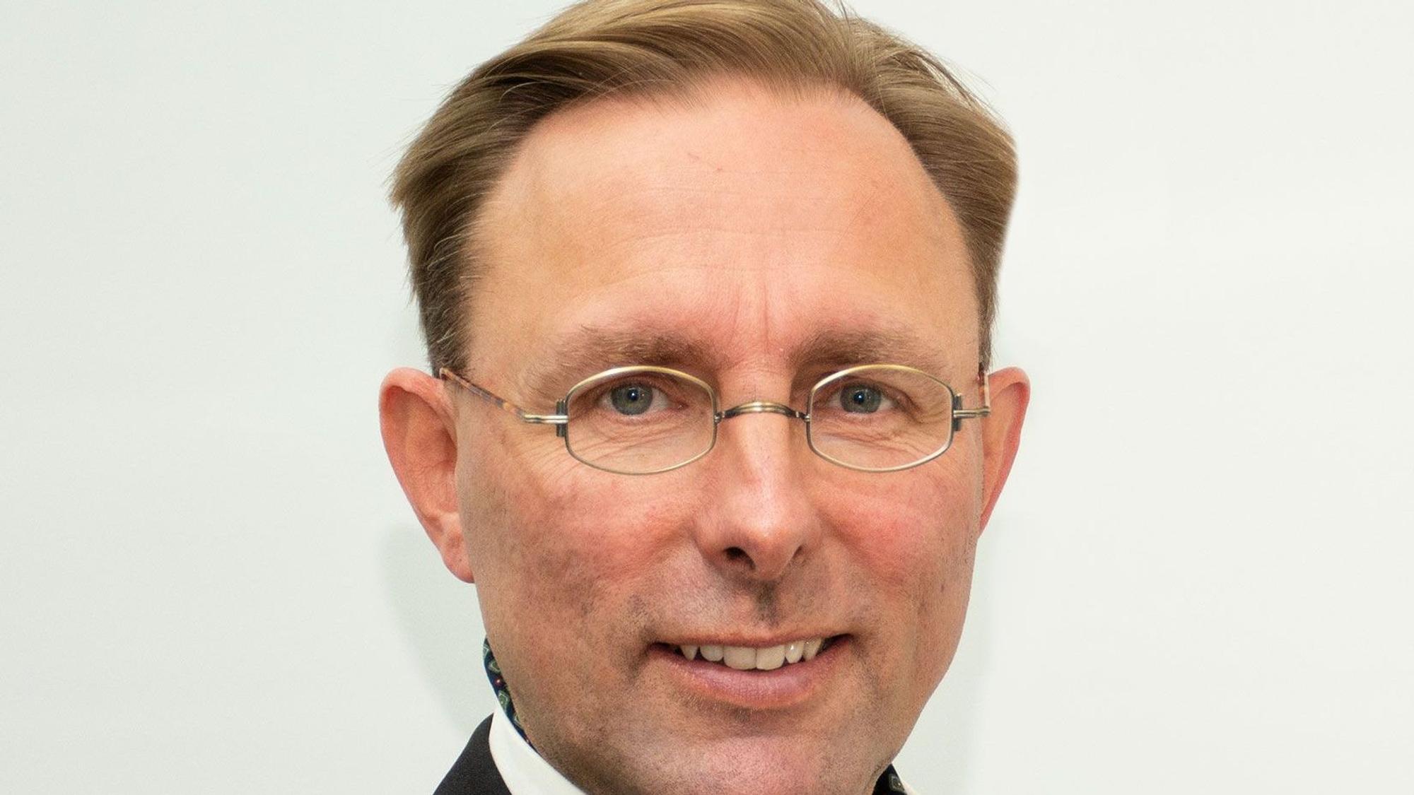 Ein Mann mit Brille im Anzug steht vor einem weißen Hintergrund und grinst.