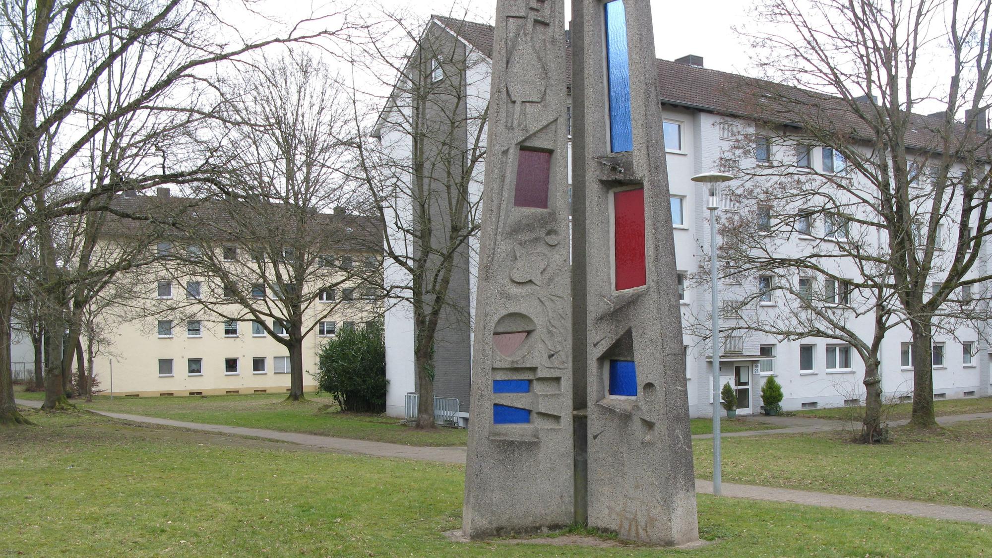 Ein Kunstwerk vor zwei Gebäuderiegeln mit Bäumen und Rasenflächen.
