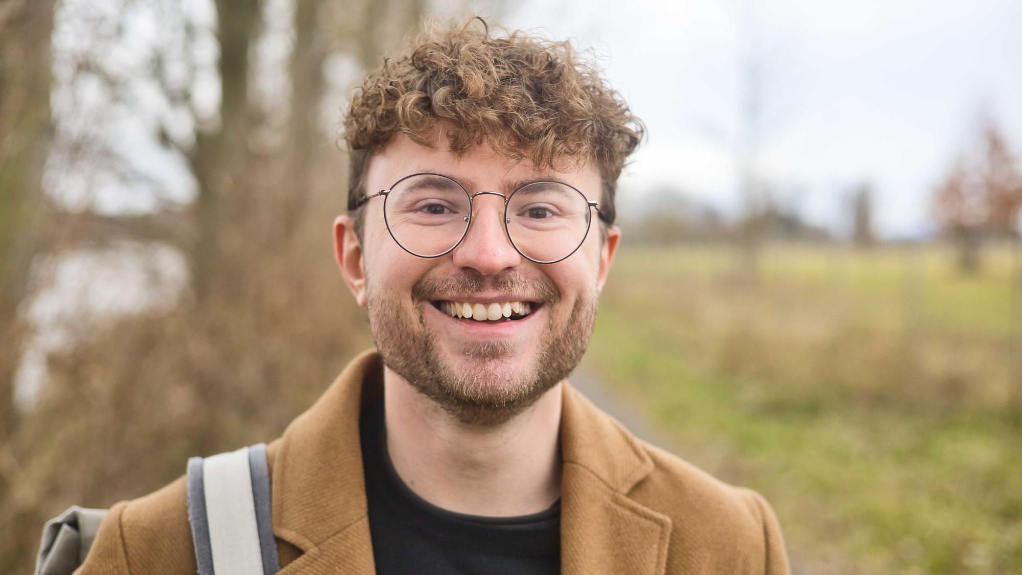 Ein Mann mit Brille steht draußen in der Natur und grinst.
