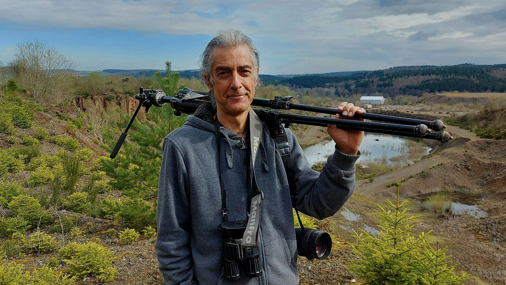 Eulen-Experte Stefan Brücher steht in der Natur. Er trägt ein Kamera-Stativ auf der Schulter.