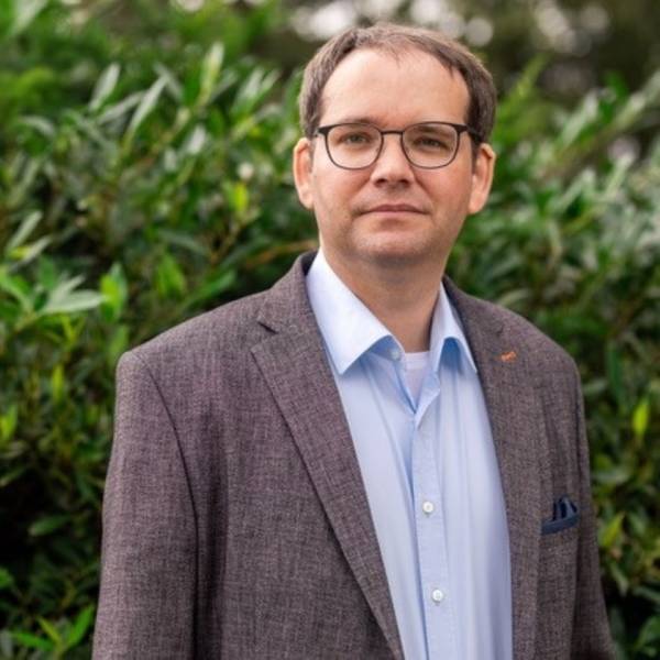 Ein Mann in Jackett, mit Brille steht vor einem grünen Busch.