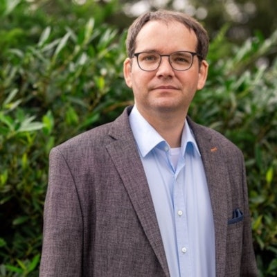 Ein Mann in Jackett, mit Brille steht vor einem grünen Busch.