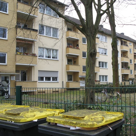 Blick über Mülltonnen und einen Zaum auf eine Hausfassade mit Balkonen, davor stehen Bäume.