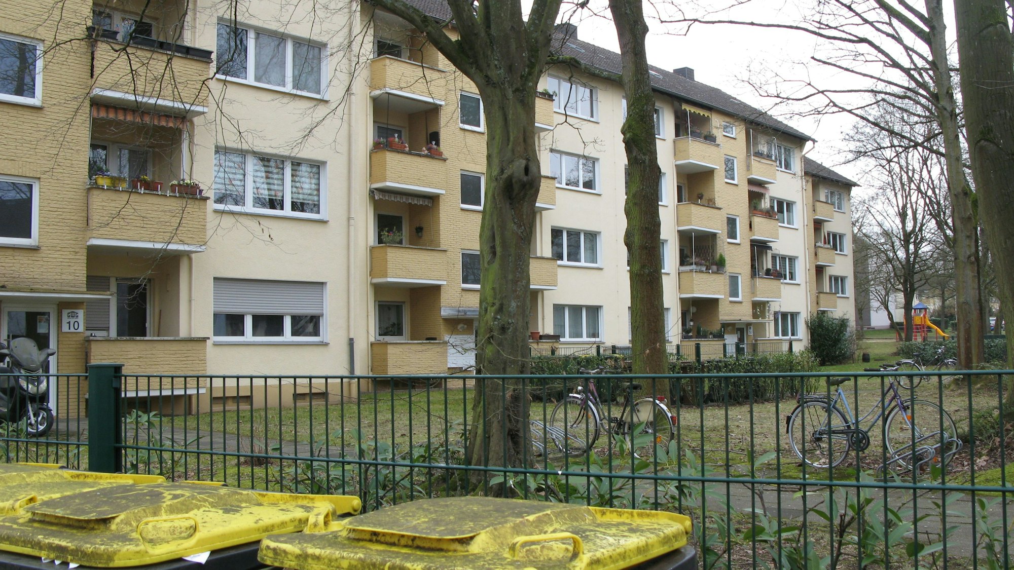 Blick über Mülltonnen und einen Zaum auf eine Hausfassade mit Balkonen, davor stehen Bäume.