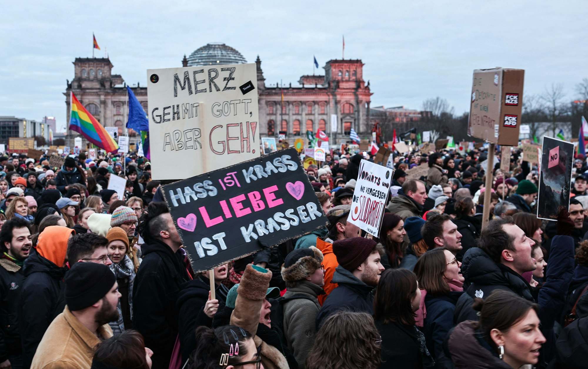 eilnehmer der Demonstration „Aufstand der Anständigen - Demo für die Brandmauer“ vor dem Reichstagsgebäude halten Plakate mit der Aufschrift „Hass ist krass Liebe ist krasser“ und „Merz geh mit Gott, aber geh!“.