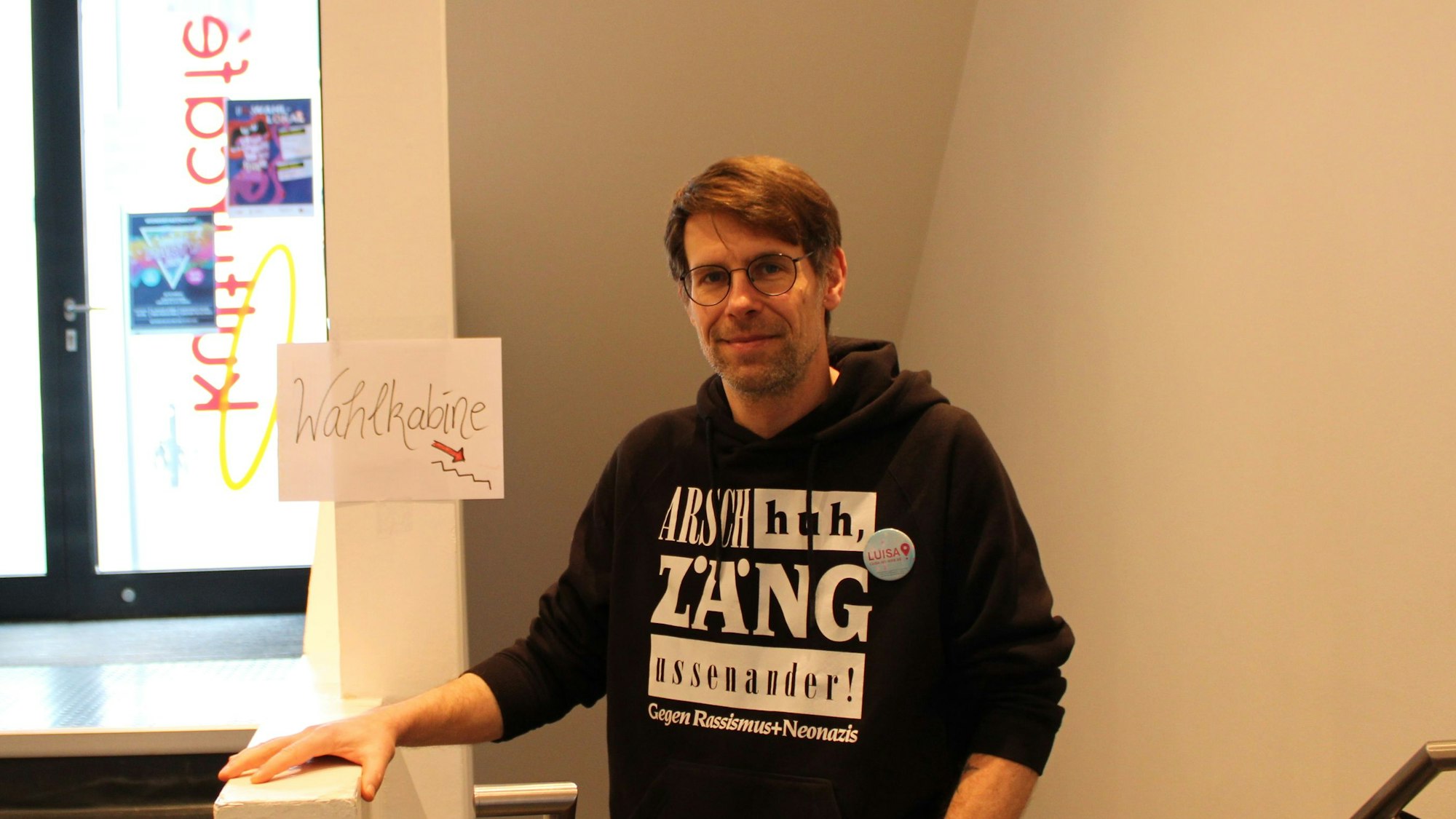 Ein Mann steht in einem Treppenhaus, daneben ist ein Schild, auf dem „Wahlkabine“ steht, ein Pfeil deutet nach unten.