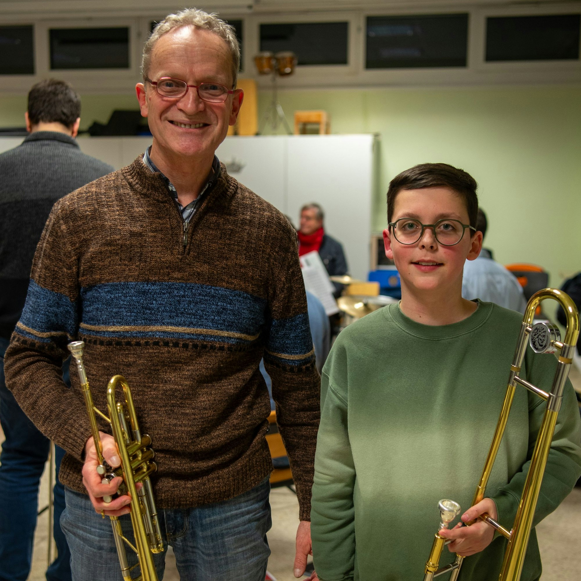 Ein Mann in braunem Pullover und mit Trompete in der Hand steht neben einem Jungen in grünem Pullover und Posaune in der Hand.