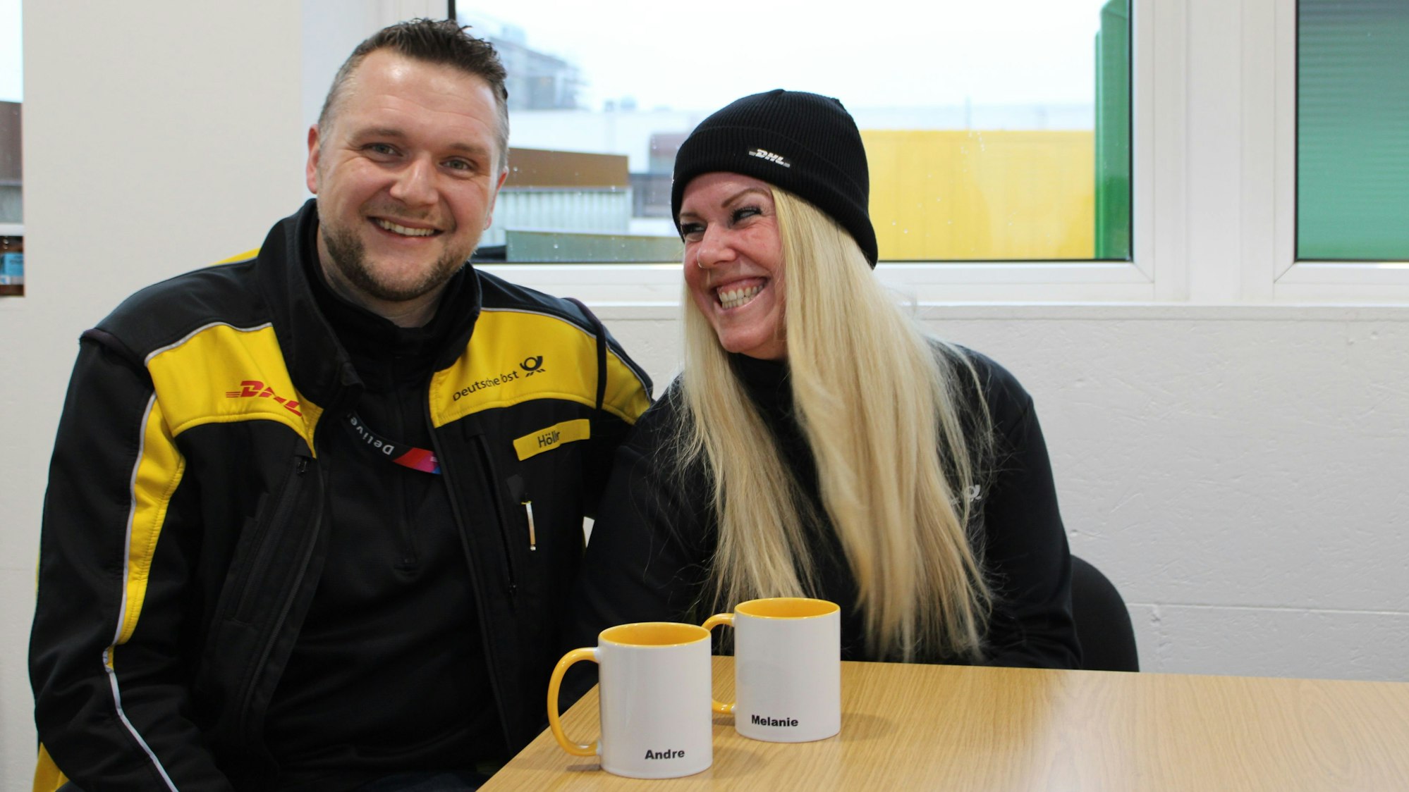 Ein Mann lächelt in die Kamera, eine Frau neben ihm schaut ihn an und lächelt breit, beide tragen ihre DHL-Kleidung. Vor ihnen stehen zwei Tassen, auf denen ihre Namen stehen: André und Melanie.
