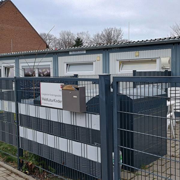 Das Bild zeigt den länglichen Container, in dem die Kita ihre Räumlichkeiten hat. Davor ein dunkelblauer Zaun.