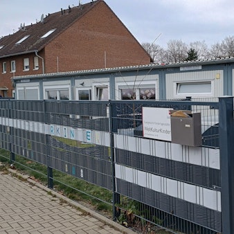 Das Bild zeigt den länglichen Container, in dem die Kita ihre Räumlichkeiten hat. Davor ein dunkelblauer Zaun.