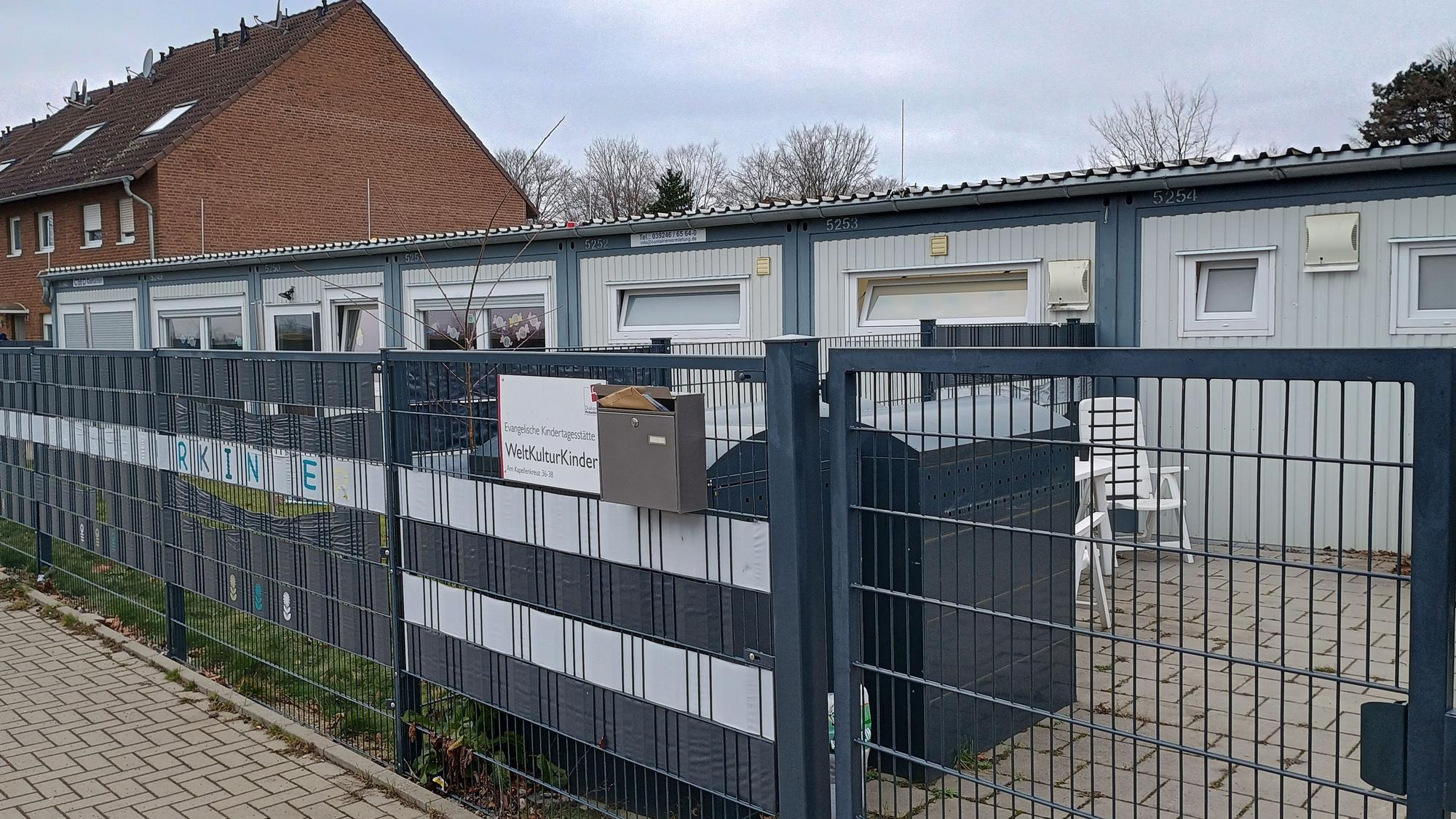 Das Bild zeigt den länglichen Container, in dem die Kita ihre Räumlichkeiten hat. Davor ein dunkelblauer Zaun.
