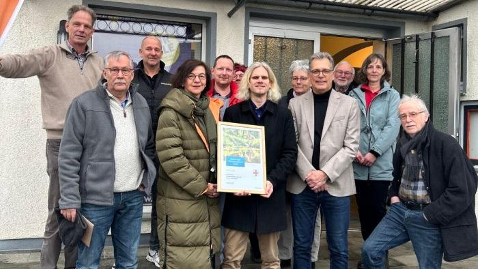Eine Gruppe Menschen steht vor einem Haus und hält eine Urkunde in die Kamera.