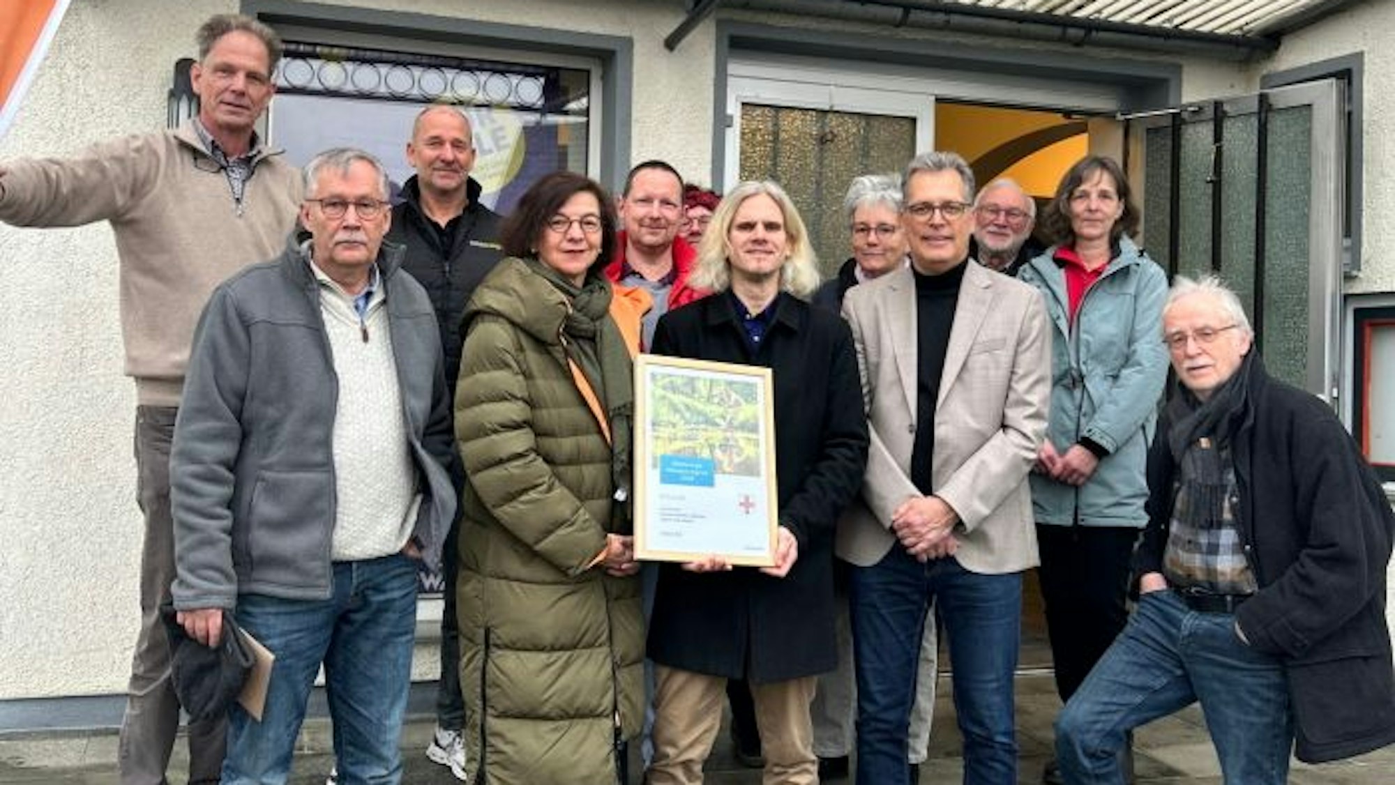 Eine Gruppe Menschen steht vor einem Haus und hält eine Urkunde in die Kamera.