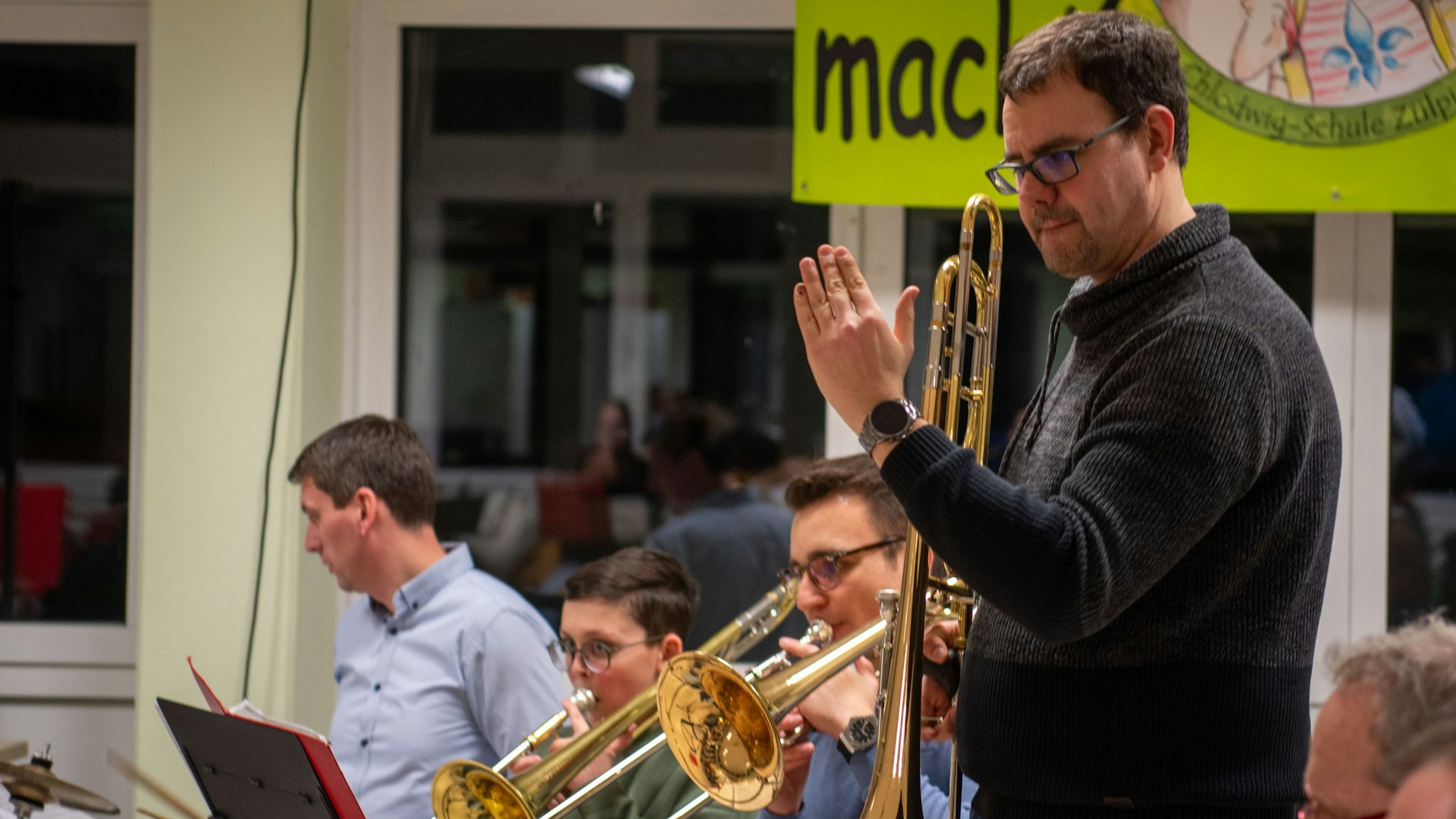 Ein Mann in dunklem Pullover steht zwischen einer Reihe Musiker und dirigiert mit Posaune in der Hand.
