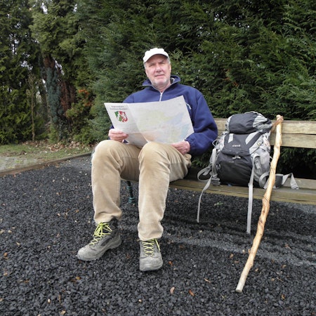 Ralf Spilles sitzt auf einer Bank. Er hält eine Karte mit dem NRW-Wappen in der Hand. Neben ihm stehen sein Wanderrucksack und ein Stock.