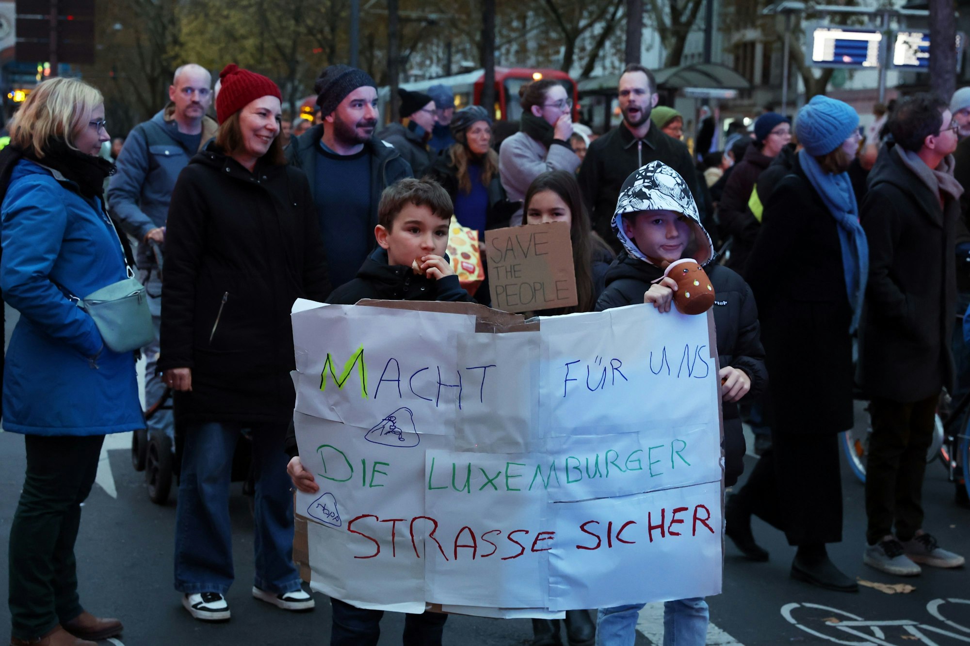 Viele Menschen sind an auf einer Straße unterwegs, zwei Kinder halten ein Plakat hoch mit dem Appell: Macht für uns die Luxemburger Straße sicher.