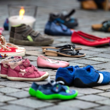 Verschiedene Schuhe von Kindern und Jugendlichen stehen auf dem Boden, dahinter ist eine Kerze zu sehen.