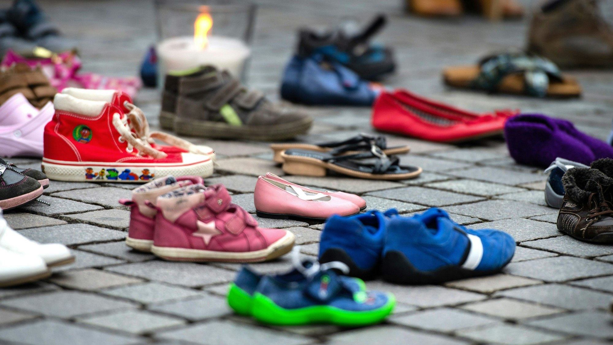 Verschiedene Schuhe von Kindern und Jugendlichen stehen auf dem Boden, dahinter ist eine Kerze zu sehen.