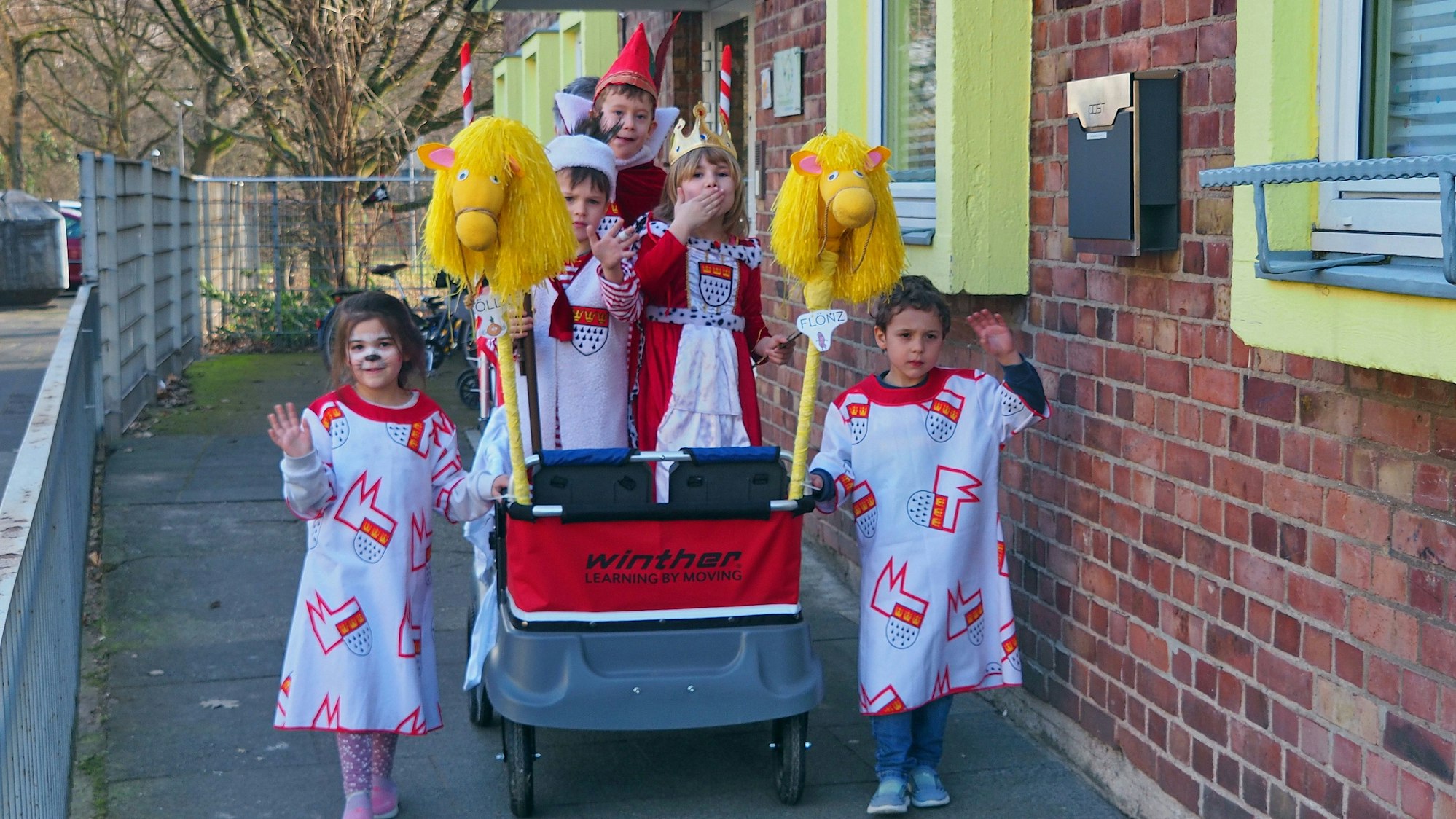 Drei Kinder stehen vor einer Kita auf einem selbst gestalteten Prinzenwagen, links und rechts gereiht von zwei Kindern als Wagenengel.