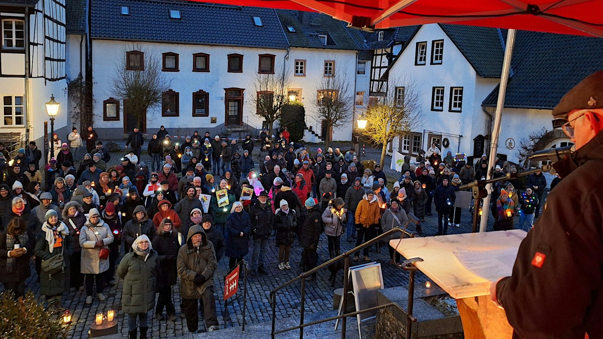 Ein Redner steht an einem Stehpult auf einer Bühne. Vor ihm auf dem Platz in Blankenheim stehen zahlreiche Menschen, von denen viele Kerzenlichter bei sich haben.