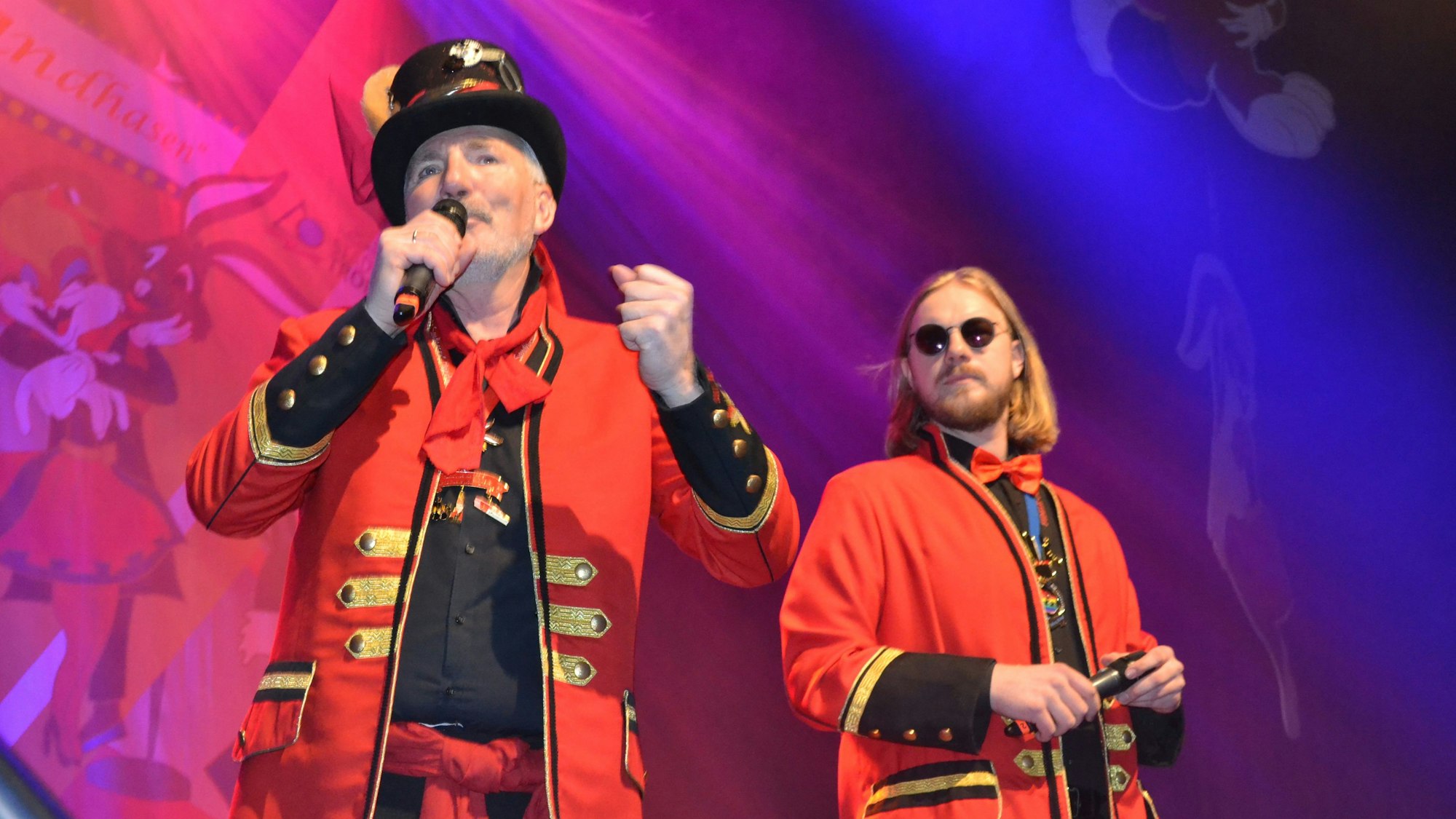 Zwei Männer in roten Uniformjacken stehen mit Mikrophonen auf einer Bühne.