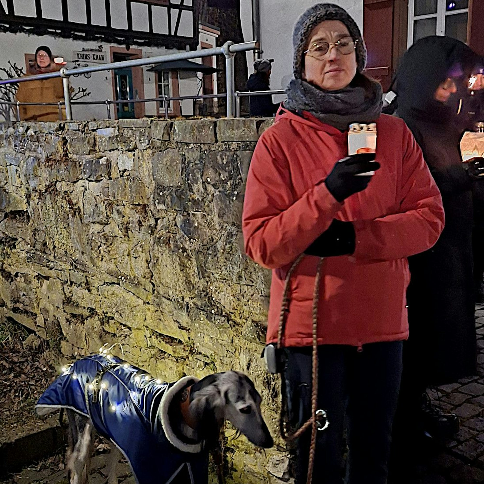 Eine Frau in roter Winterjacke hält ein Windlicht in der Hand. An der Leine führt sie ihren Hund, dessen Hundemantel mit einer Lichterkette geschmückt ist.