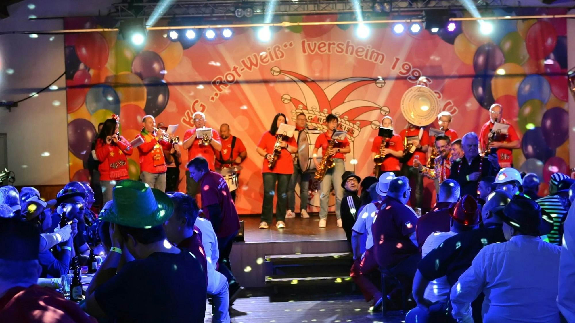 Musiker in roten Shirts stehen auf einer Bühne und spielen. Die Menschen in Saal sind in blauem Licht angestrahlt.