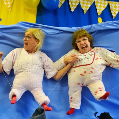 Drei der Jecken Wiewer haben sich in Babys verwandelt: Ihre Köpfe und Arme haben sie durch ein blaues Tuch gesteckt, auf dem kleine ausgestopfte Strampler angebracht sind.