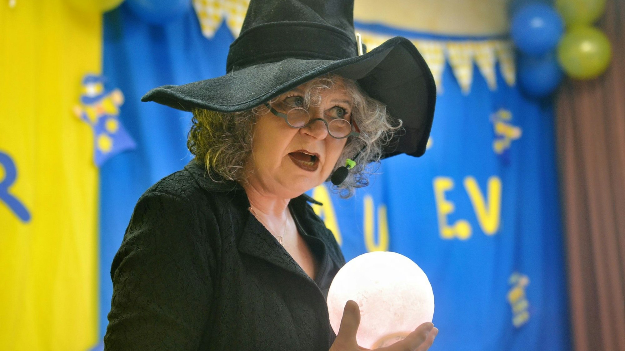 Eine Frau mit grauen Locken und einem schwarzen Hexenhut hat eine leuchtende Glaskugel in der Hand. Im Hintergrund sieht man die blau-gelbe Dekoration der Jecken Wiever in der Alten Schule.