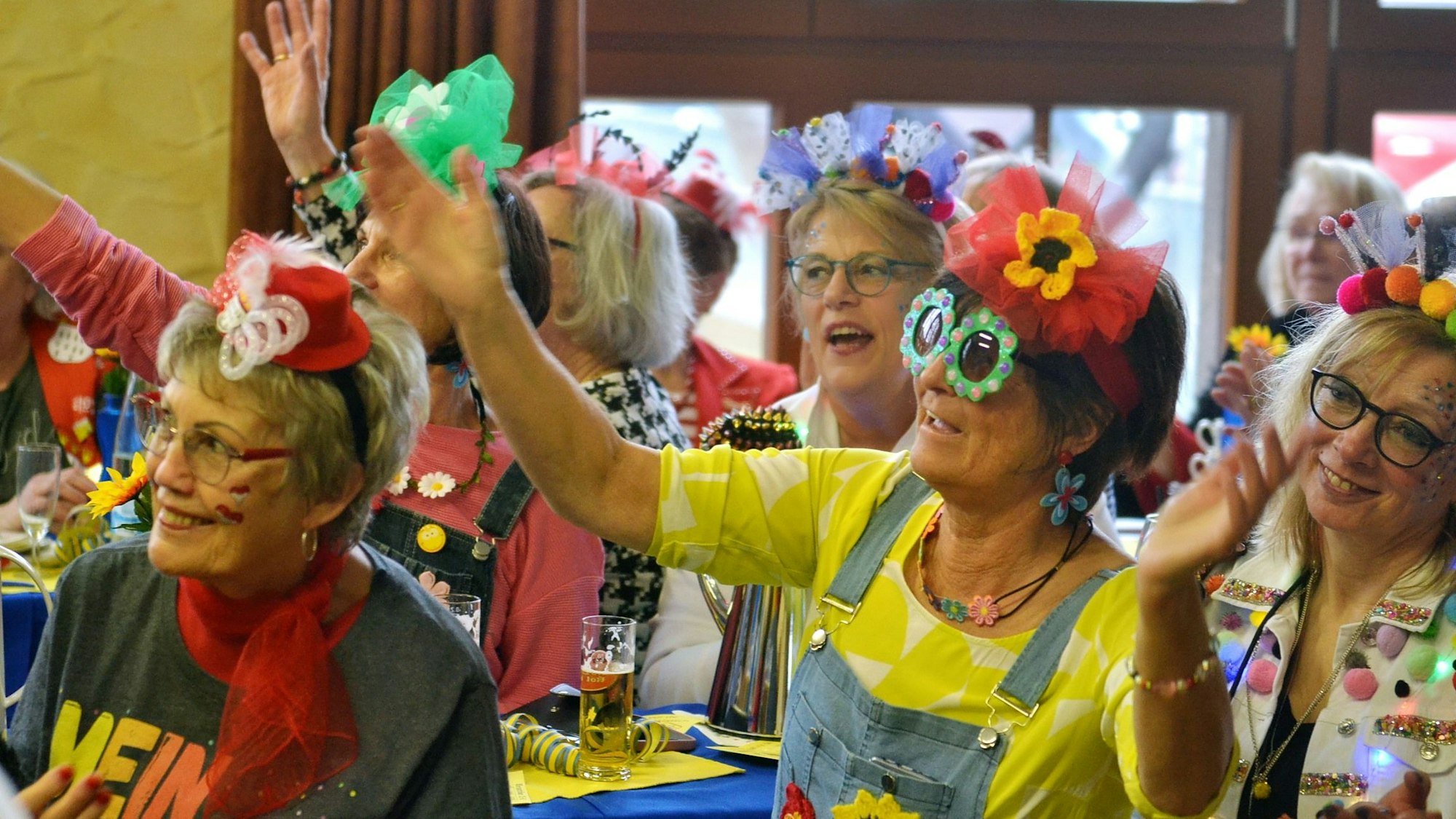 Ausgelassen singende und winkende Antweiler Närrinnen, die lustig kostümiert sind, sitzen bei der Dorfsitzung zusammen und schauen auf die Bühne.