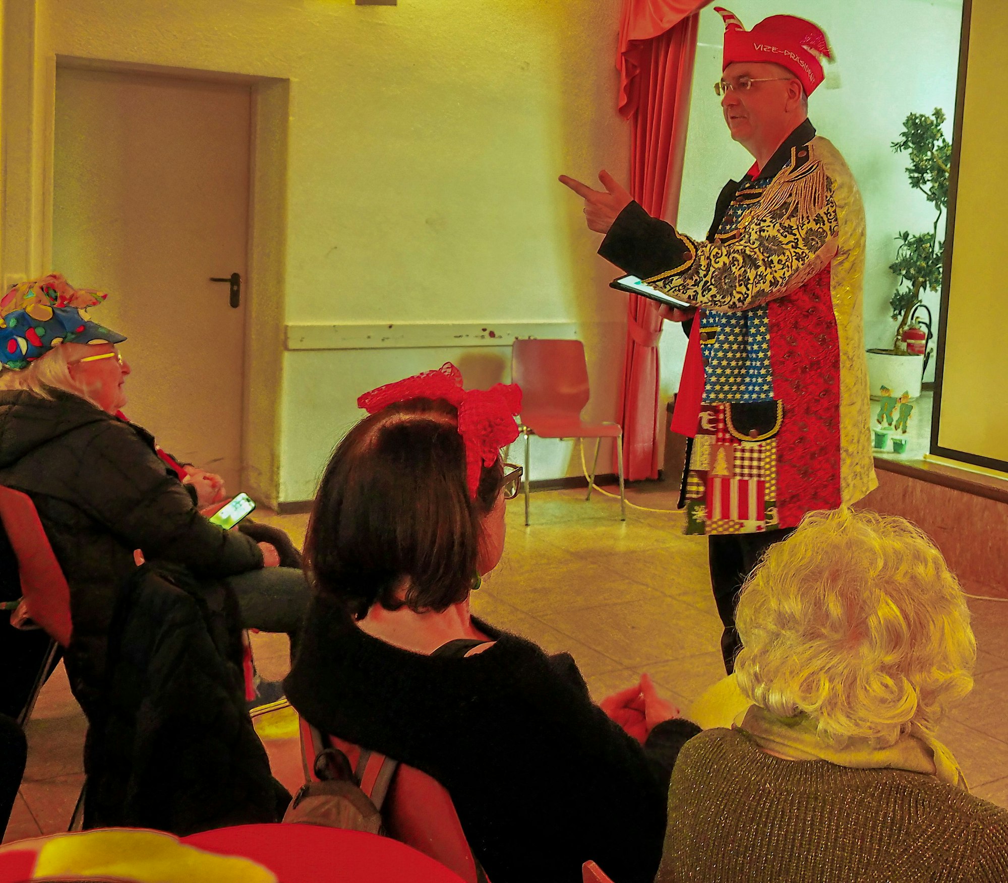 Ein Mann mit Narrenkappe und bunter Anzugjacke steht vor gefüllten Sitzreihen in einem Saal und gestikuliert.